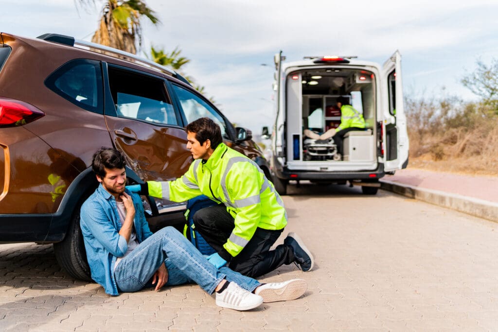 A paramedic wearing a neon jacket and gloves attends to a young injured man sitting on the ground near a brown SUV.