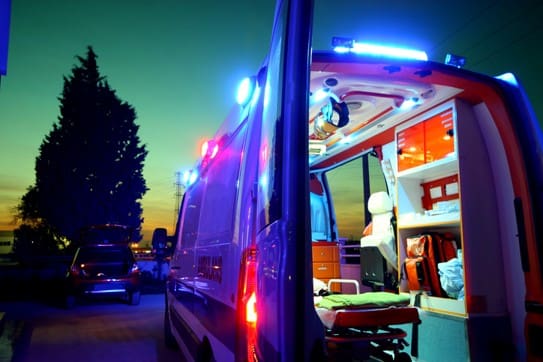An ambulance with its back door open under a twilight sky. Emergency lights illuminate the scene, revealing medical equipment inside, creating a sense of urgency.